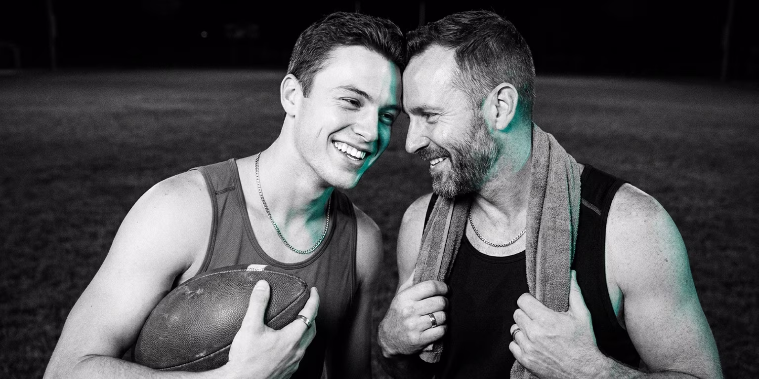 Two teammates grinning at each other on a sports field at night, one holding a football, lit by a teal light